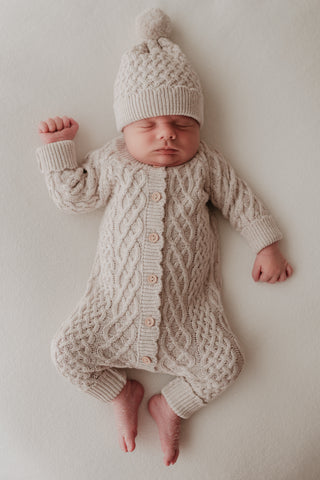 Sleeping newborn wearing light grey cable knit romper and pom pom hat