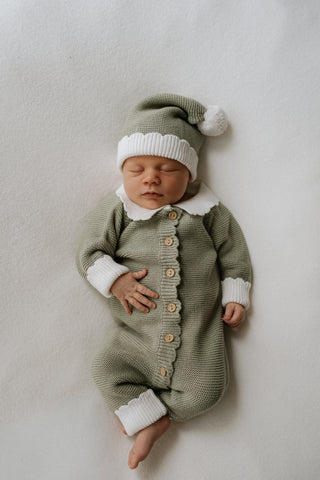 Baby wearing a sage green knitted Christmas Elf romper and hat with white collar, cuffs, and pom pom