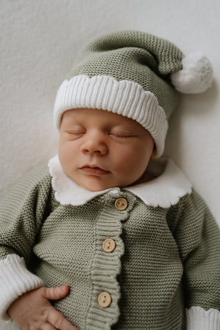 Baby wearing a sage green knitted Christmas Elf romper and hat with white collar, cuffs, and pom pom
