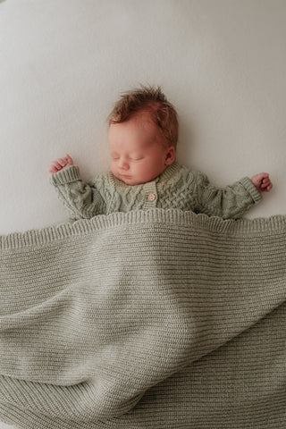 Baby wearing sage green cable knit cardigan and sleeping under a sage green scalloped edge blanket
