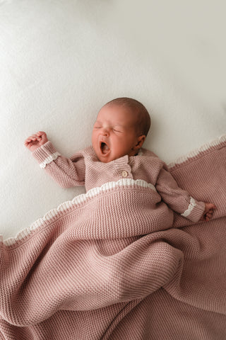 Yawning baby wearing pink knitted romper with a matching pink knitted baby blanket with a cream scalloped trim 