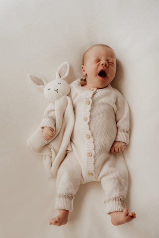 Yawning baby wearing a cream knit romper with a matching bunny comforter