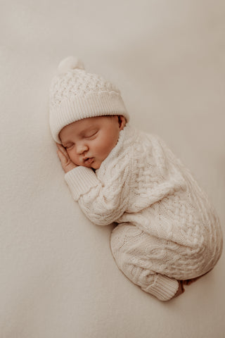 Sleeping baby lying on their front wearing cream cable knit romper and hat