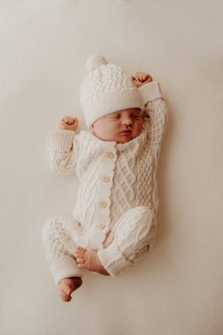 Stretching baby wearing cream cable knit romper and hat