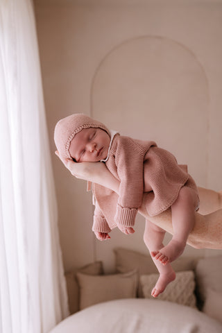 Sleeping newborn wearing a pink knitted outfit being held up near a white curtain