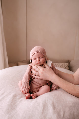 Sitting baby wearing a pink knitted outfit and bonnet being supported by mother's hands