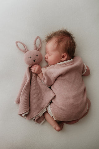 Baby wearing pink knit bodysuit with cream scalloped edge cuddling pink bunny comforter