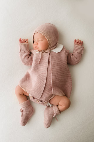 Sleeping baby wearing pink knitted bonnet, cardigan, and booties