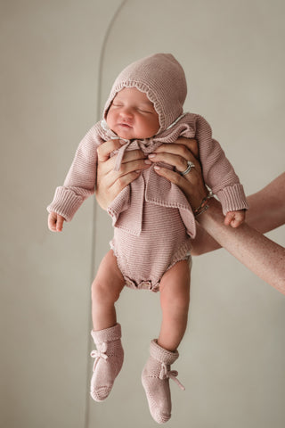 Baby being held up wearing a pink knitted bonnet. cardigan, romper, and booties 