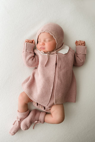 Sleeping baby wearing a pink knitted bonnet, booties, and cardigan with white collar 
