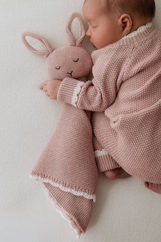 Sleeping baby wearing pink knitted romper cuddling a pink knitted bunny comforter 