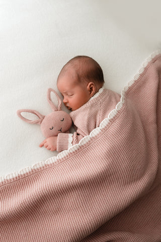 Sleeping baby wearing a pink knitted outfit, with a pink knitted bunny comforter with cream ears, and a pink knitted blanket, all finished with cream contrast scalloped edges. 