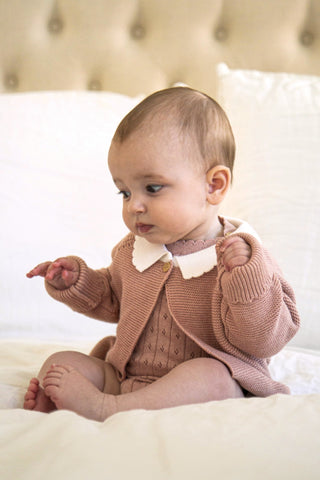 Baby sitting on a bed wearing a pink knitted cardigan with white collar