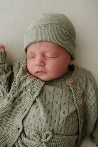 Sleeping baby wearing sage green knit hat, bodysuit and cardigan