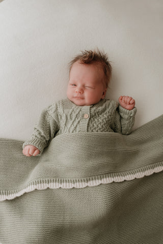 Baby wearing a sage green cable knit cardigan with a matching sage green blanket with a cream scalloped edge