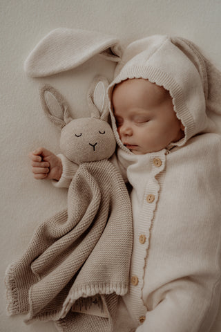 Baby wearing a knitted bunny ear romper cuddling a grey knitted bunny comforter