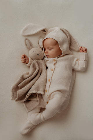Baby wearing a knitted bunny ear romper cuddling a grey knitted bunny comforter