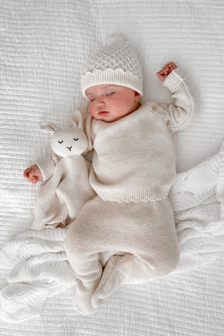 Sleeping baby lying on white quilted bedding wearing a beige knitted outfit and hat, cuddling a bunny comforter