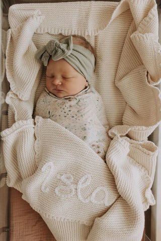 Sleeping newborn baby with sage green bow headband, floral swaddle, and knitted embroidered 'Isla' blanket