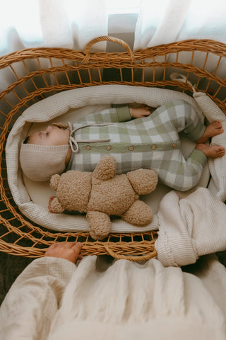 Sleeping baby in moses basket wearing sage gingham knitted romper and beige bonnet cuddling a teddy bear