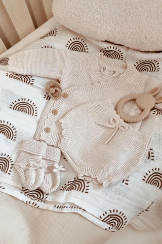 Knitted beige baby outfit laid out on rainbow muslin with dummy and teether