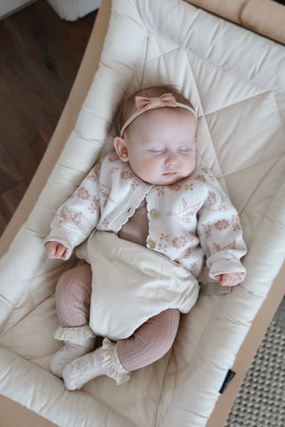 Sleeping baby in rocker chair wearing pink bow headband, knitted pattern cardigan, pink leggings, and frilly socks