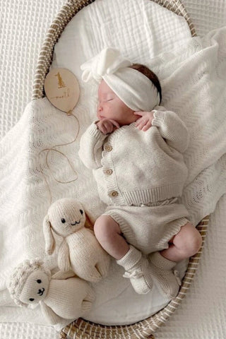 Baby in basket, wearing bow headband, and knitted outfit, with white blanket, 1 month wooden balloon plaque and knitted toys. 