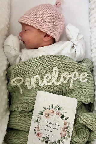 Sleeping newborn baby wearing pink knitted hat and with sage embroidered 'Penelope' blanket with baby name announcement card