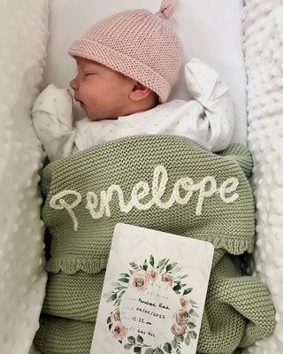 Sleeping newborn baby wearing pink knitted hat and with sage embroidered 'Penelope' blanket with baby name announcement card