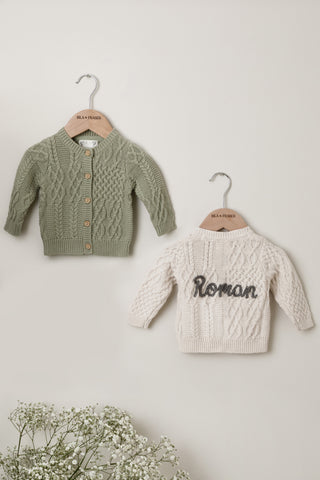 Pair of cable knit Baby Cardigans on Isla & Fraser hangers, one in sage green and one in beige with 'Roman' name embroidery. 