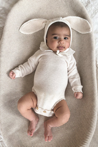Baby in basket lying on beige blanket wearing a beige bunny ear bonnet and bodysuit with frill collar